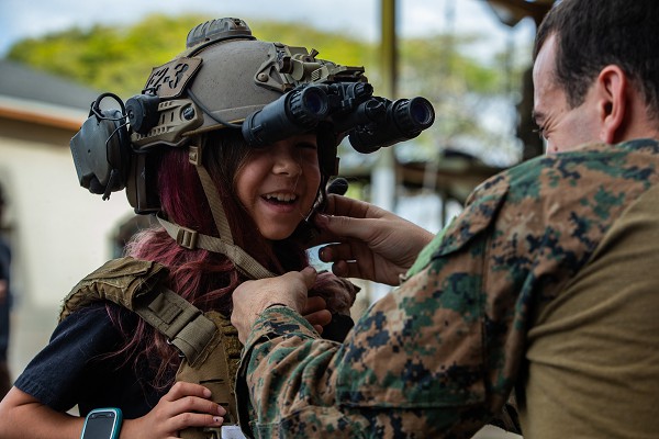 In this U.S. Marine Corps photo by Corporal Isaac Cantrell, a “military brat” is fitted with a helmet during a tactical demonstration/community relations event at Kapolei, Hawaii.  Service kids like these endure the instability and emotional separations common to military life.