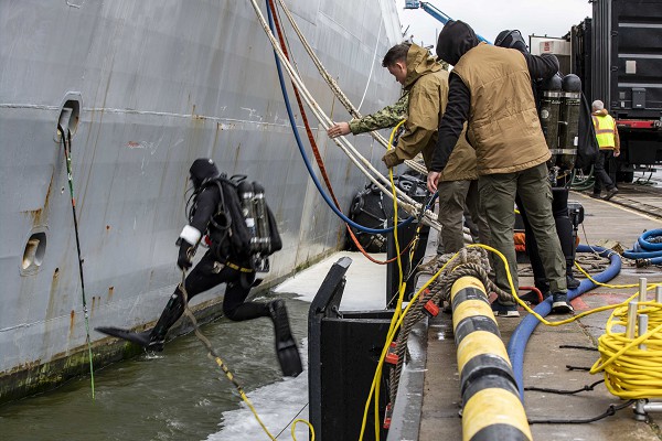 Klaipeda, Lithuania (November 19, 2022): In this photo by MC1 John Bellino, U.S. Navy Diver 1st Class William Crampton, left, assigned to Forward Deployed Regional Maintenance Center, Bahrain, enters the water to conduct underwater maintenance on the San Antonio-class amphibious transport dock ship USS Arlington during a scheduled port visit. The Arlington is part of the Kearsarge Amphibious Ready Group and 22nd Marine Expeditionary Unit, under the command and control of Task Force 61/2, is on a scheduled deployment in the U.S. Naval Forces Europe area of operations, employed by U.S. Sixth Fleet to defend U.S., allied and partner interests.